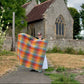 A person outdoors in front of a stone church holding up a large, colorful plaid quilt or blanket spread wide to show its pattern. The quilt features a multicolor plaid design with shades of orange, yellow, red, green, blue, and purple.
